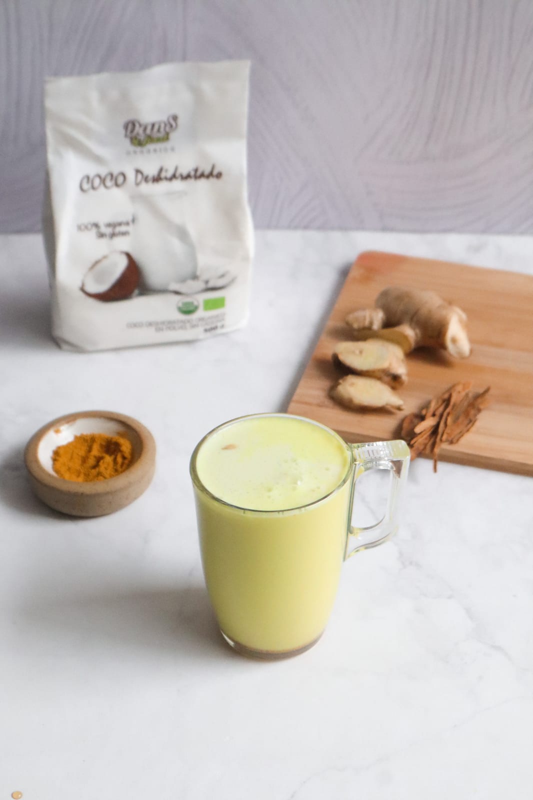 Taza de leche dorada espumosa sobre mármol con cúrcuma, jengibre fresco y canela en astillas